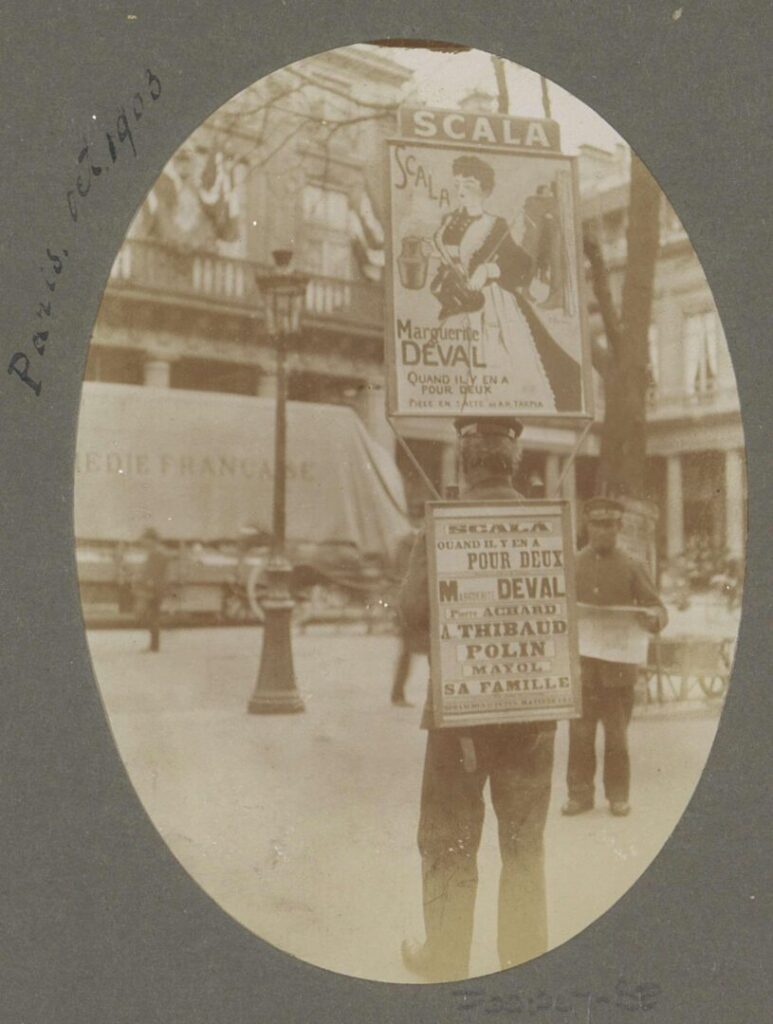 A sepia photograph in a grey oval cardboard frame. On the frame handwriting in ink specifies "Paris, Oct. 1903". The image shows a Parisian street in the background and in the foreground a man wearing a suit and hat with his back to the camera, wearing one large sign over his torso and an additional one above his head. The two signs are advertising a theater piece with Marguerite Deval at Scala.