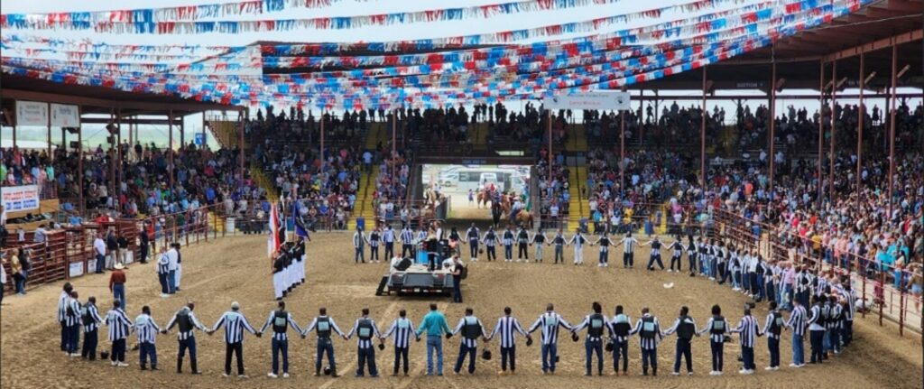 A large group of incarcerated participants standing in a circle and holding hands in the center of the arena during the opening ceremony before a seated audience.