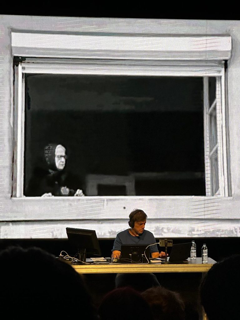 An old woman at a window appears in a black and white projection. In front of it, a man wearing headphones sits at a desk with computers.