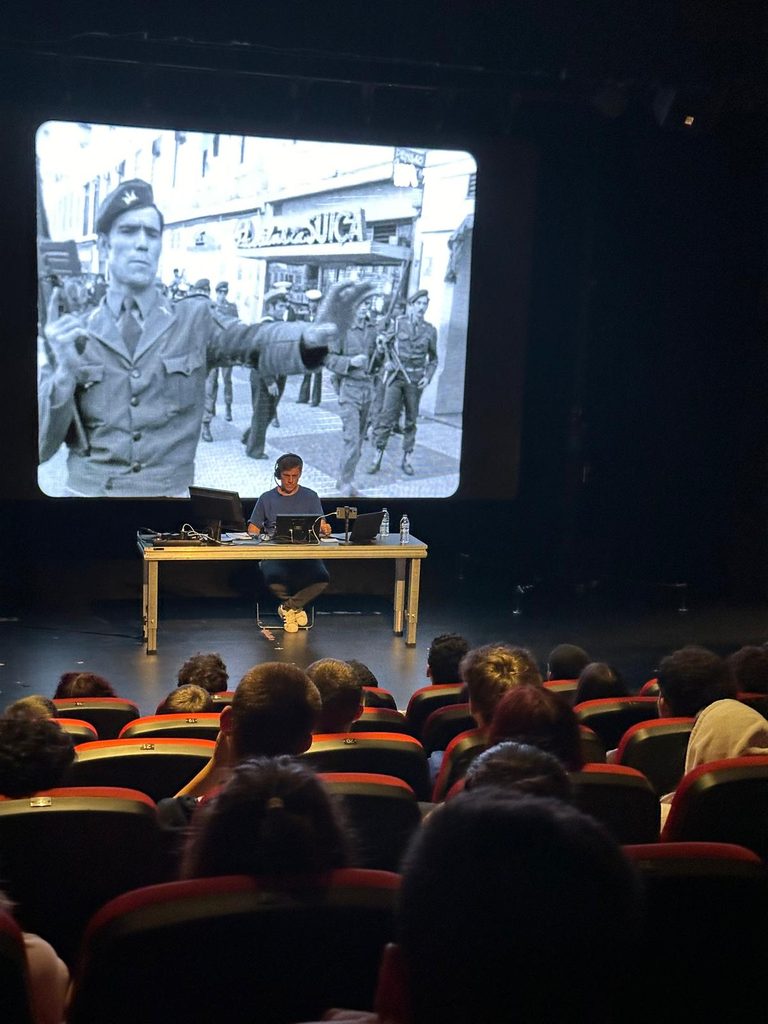 Soldiers in the streets appear in a black and white projection. In front of it, a man a man wearing headphones sits at a desk with computers. People siting in a theatre auditorium look a them.
