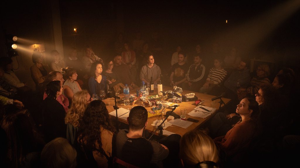 At the center of the photo, there is a table with bottles of water, wine, and used snack packages. Many people are sitting around the table in concentric rows. One cannot distinguish between the actors and the spectators, except that a few people have mics.