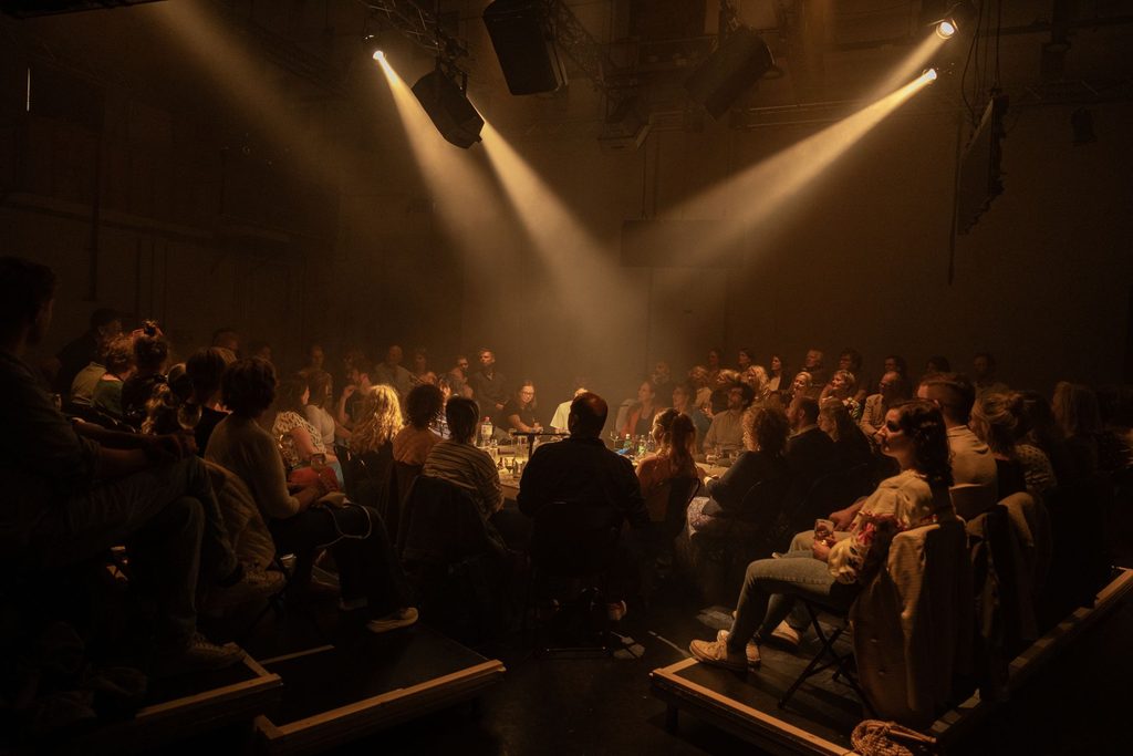 At the center of the photo, there is a table with bottles of water, wine, and used snack packages. Many people are sitting around the table in concentric rows. One cannot distinguish between the actors and the spectators, except that a few people have mics.