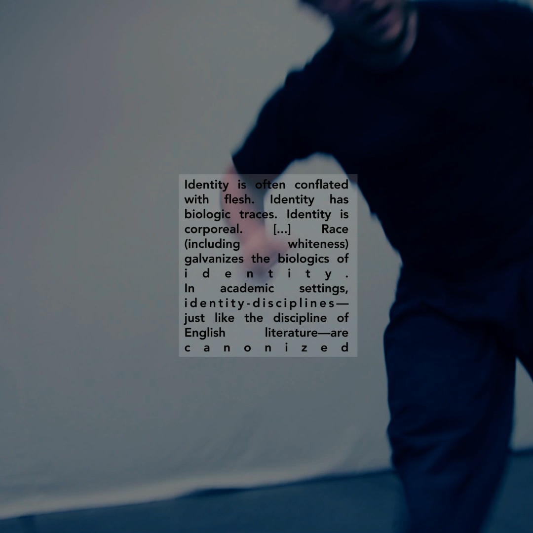 An apparently white figure moves in an empty studio, partially out of frame. Colorshift blue. Overlaid text on race, identity, whiteness, and disciplinary.