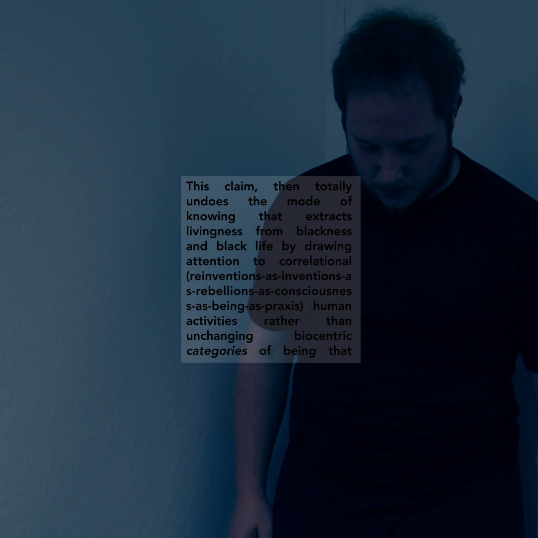 An apparently white figure stands in an empty studio. Colorshift blue. Overlaid text on undoing modes of knowledge that extract livingness from blackness.